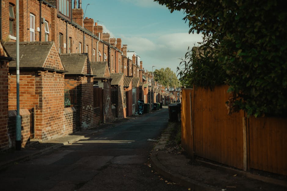 A quiet residential street scene during daytime with a row of brick terraced houses on the left side, each featuring small front yards with brick walls and steps leading up to their doors. The houses have pitched roofs with chimney stacks, and the facades are constructed from red brick. On the right side, a tall wooden fence runs along the pavement, partially obscuring greenery and bushes behind it. The street is narrow, with a black waste bin positioned near the curb and a few other bins visible further down the alley. The ground is paved with asphalt, showing slight wear and patchwork repairs. Shadows cast by the buildings suggest the sun is low in the sky, possibly in early morning or late afternoon. This scene reflects a typical urban neighborhood, often involved in home relocations or furniture transport, where careful planning and logistics are essential in the process of packing and moving household items, which local removals specialists like Man and Van Mitcham assist with efficiently.