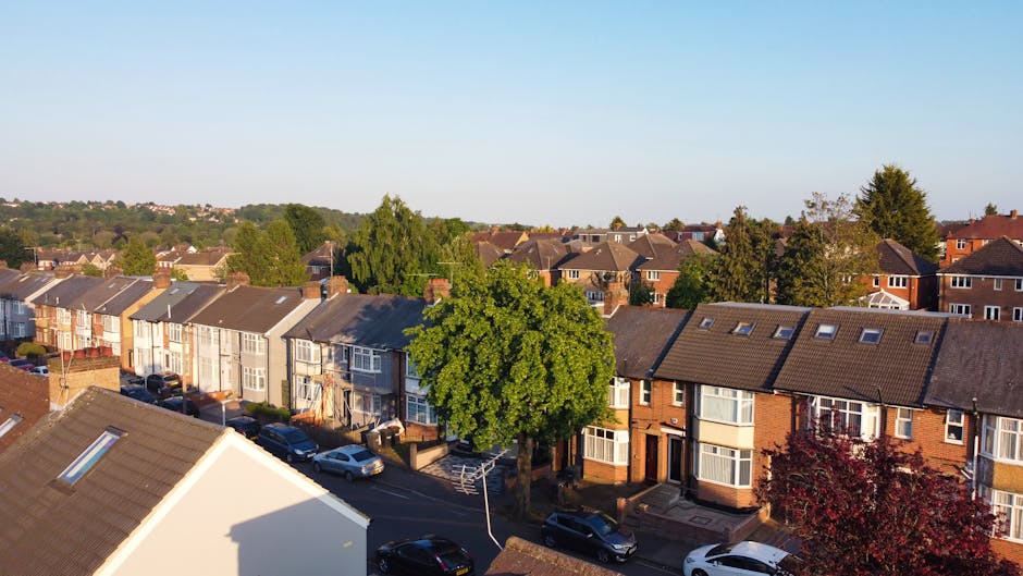 An aerial view of a residential street in Mitcham during daylight hours, showing a row of terraced houses with uniform rooftops, some with skylights, and a mix of brick and painted exteriors. A large, leafy green tree is centered along the pavement, casting shadows onto the parked cars lining the road. Several vehicles, including vans, cars, and possibly a small moving truck, are parked along the curb, indicating activity related to home relocation or furniture transport. In the foreground, part of a house's roof with a skylight is visible, and the street appears quiet with no visible people. The environment includes well-maintained front gardens, paved walkways, and some houses with bay windows or small front porches, all set under a clear blue sky. This scene exemplifies a typical residential area where professional removals services, such as those offered by Man and Van Mitcham, might assist with packing, loading, and transporting household items during a home relocation process.