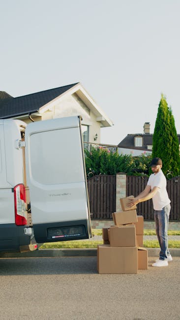 A man wearing a white t-shirt and jeans is standing on the pavement beside a silver van with its rear doors open, loading and stacking cardboard boxes of varying sizes. The boxes are arranged on the ground and in the process of being transferred into the van, which is parked close to a residential property with a white exterior and dark roof. The scene takes place outdoors during daylight, with a clear sky visible above, and features a garden with green bushes and a tall, conical evergreen tree behind a wooden fence. The man appears to be involved in packing or moving items as part of a home relocation or furniture transport service, with protective packaging materials such as plastic wrap and cardboard boxes contributing to the packing and moving process. This image is associated with house removal services provided by Man and Van Mitcham, supporting attentive relocation logistics within the context of house removals.