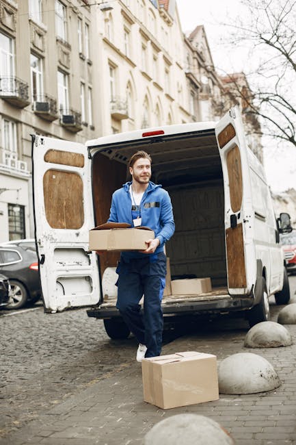 A man wearing a white t-shirt and jeans is standing on the pavement beside a silver van with its rear doors open, loading and stacking cardboard boxes of varying sizes. The boxes are arranged on the ground and in the process of being transferred into the van, which is parked close to a residential property with a white exterior and dark roof. The scene takes place outdoors during daylight, with a clear sky visible above, and features a garden with green bushes and a tall, conical evergreen tree behind a wooden fence. The man appears to be involved in packing or moving items as part of a home relocation or furniture transport service, with protective packaging materials such as plastic wrap and cardboard boxes contributing to the packing and moving process. This image is associated with house removal services provided by Man and Van Mitcham, supporting attentive relocation logistics within the context of house removals.