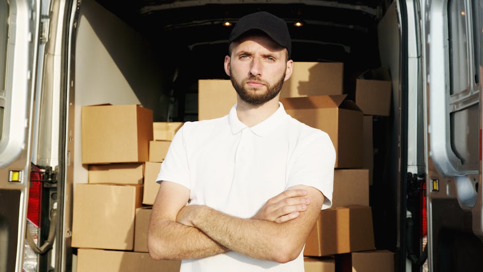 A young man with a beard and wearing a black cap and white polo shirt stands inside the back of a van used for house removals, with his arms crossed and a serious expression. Behind him, the van's interior is filled with neatly stacked cardboard boxes of various sizes, some sealed with packing tape, ready for a home relocation. The van's open rear doors reveal a paved area outside, suggesting the loading or unloading process is underway. The environment is well-lit, with natural light illuminating the scene. This image illustrates the practical aspects of furniture transport, packing, and loading during professional removals services, as provided by Man and Van Mitcham, highlighting the logistics involved in household moves.