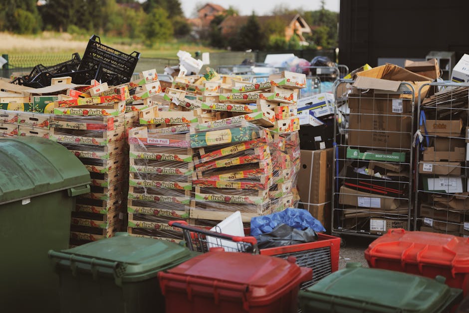 An outdoor scene showing multiple stacks of cardboard boxes filled with fresh fruits, arranged on wooden pallets on a paved surface, with some black plastic crates stacked next to them. Several large plastic waste bins with closed lids in green and red are positioned in the foreground. To the right, a metal trolley with cardboard boxes and packaging materials is visible, along with various smaller boxes and plastic bags scattered around. The background includes a residential area with houses and greenery, under natural daylight conditions. This image reflects the logistics involved in home relocation, specifically the packing and loading process, as handled by Man and Van Mitcham during furniture transport or household moves, consistent with the service information related to house removals and bulky waste disposal outlined on the Mitcham relocation guidelines page.