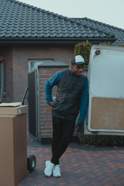 A man wearing a blue beanie, a navy and red windbreaker, black pants, and white sneakers sits on the edge of an open van with its sliding door open. Inside the van, several large cardboard boxes are s