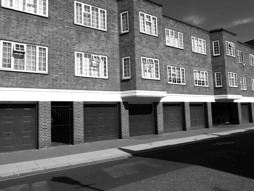 A black and white photograph of a modern, multi-storey residential building with a brick exterior, featuring multiple rows of white-framed windows, some of which are open. The ground level has several