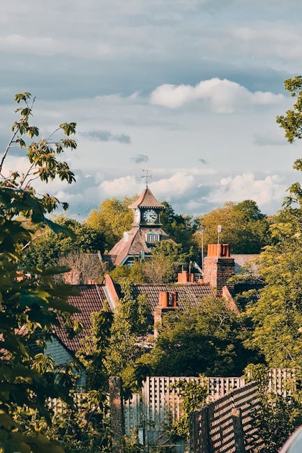 A scenic view of a residential area with a prominent historic clock tower topped with a copper-colored, pointed roof in the distance, surrounded by a variety of green trees with dense foliage. The foreground features rooftops of houses with red and brown tiles, some chimney stacks, and a wooden fence partially visible at the bottom. The scene is captured during daylight with partly cloudy skies, showcasing natural light illuminating the buildings and greenery. This setting reflects a typical UK suburban environment that might be involved in home relocation activities, where careful packing, loading, and transport of household items could take place, supported by professional removals services such as Man and Van Mitcham. The image emphasizes both the scenic beauty of the area and the context of house moving logistics nearby Mitcham Common.