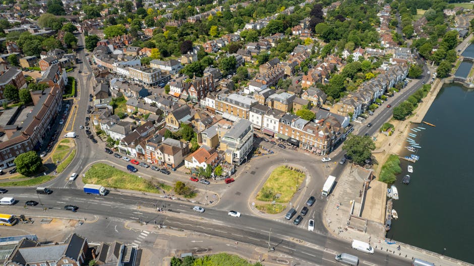 An aerial view of a residential area near Mitcham Common showcasing a mix of terraced and semi-detached houses with brick facades, pitched roofs, and front gardens. The streets are lined with parked cars and move with a flow of vehicles, including vans and small trucks. A roundabout at the intersection is visible with a few vehicles navigating around it. To the right, a river runs alongside a promenade with boats moored along the water’s edge. The surrounding landscape features lush green trees and well-maintained lawns, indicating a quiet neighborhood setting ideal for house removals. The image captures a sunny day with clear weather, highlighting the urban layout and proximity to natural spaces, supporting the context of home relocation and furniture transport, as managed by companies like Man and Van Mitcham during packing and loading processes.
