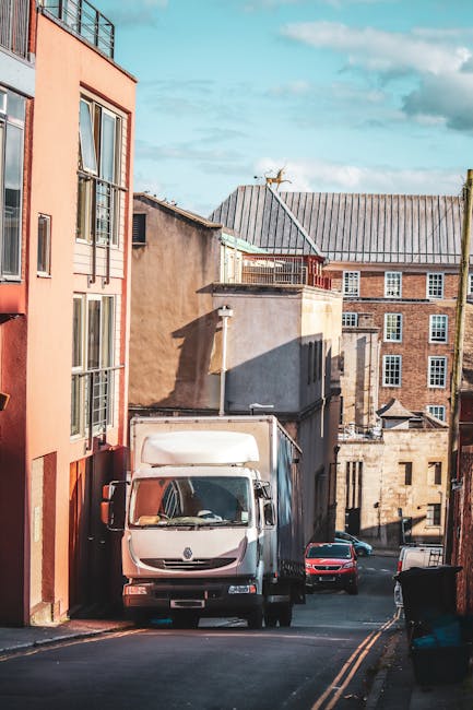 A white moving van parked on a narrow urban street with residential buildings lining both sides. The van is positioned close to the pavement, with its front facing slightly right. To the left, a multistory property with large windows and orange exterior walls is visible, partially casting shadow on the street. Behind the van, there are other vehicles, including a red car, and various tones of brick and concrete buildings with flat roofs and small windows. The setting suggests a typical home relocation scenario in a tight streetscape, where [COMPANY_NAME] might undertake furniture transport and packing and moving services through challenging narrow access routes, with efforts likely underway to load or unload belongings. The sky above is partly cloudy, and the overall scene highlights the logistics involved in removals within confined city environments.