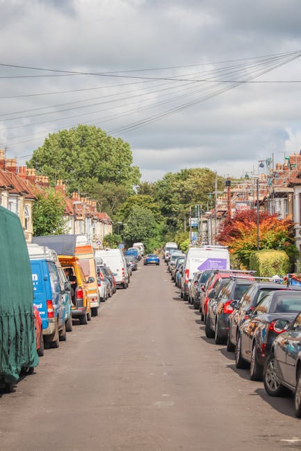 A residential street with parked cars lining both sides of the narrow asphalt road, including various makes and models covered with protective fabric and plastic wrap used during home relocation. In the background, green trees and a partly cloudy sky are visible above rows of terraced houses with brick facades, small front gardens, and chimneys. Several moving boxes and pieces of furniture, such as a wardrobe and cardboard cartons, are placed near the houses' doorways, indicating ongoing packing or loading activities. A man and van service, Man and Van Mitcham, is likely involved in the furniture transport and packing process, supporting house removals with narrow access challenges. The scene suggests careful loading and transportation of household items through a limited-access residential street as part of a professional moving service. The surroundings are well-lit with natural daylight, emphasizing the careful handling of items within this relocating process.