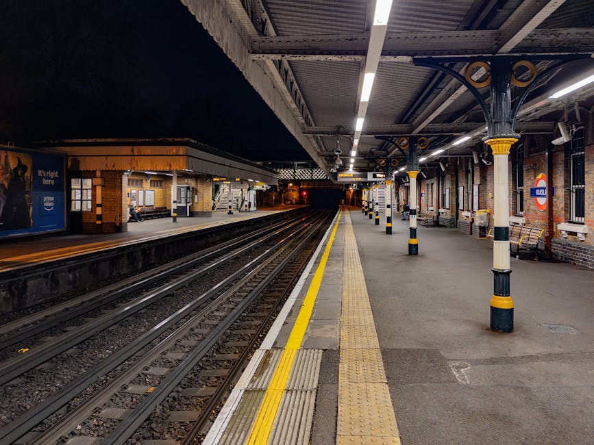 A well-lit, empty train platform at night with multiple sets of railway tracks running parallel, separated by a yellow safety line along the edge. The platform features brick walls with large windows and a roundel sign indicating the station's name. Support columns with decorative yellow and black bands line the platform, alongside benches and informational signage. The station's overhead roof, supported by metal beams, extends across the platform, and the lighting creates a clear view of the area. This scene depicts a typical UK train station environment suitable for home relocation, furniture transport, or packing and moving services, with the platform prepared for the arrival or departure of trains, as managed by providers like Man and Van Mitcham.