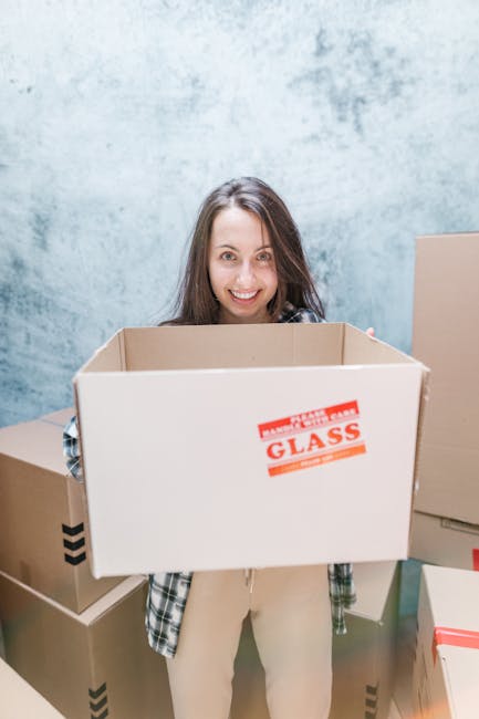 A person dressed in a dark-colored shirt and blue pants is shown in a close-up image during a home relocation process, holding a cardboard box sealed with packing tape. The individual is affixing a wh
